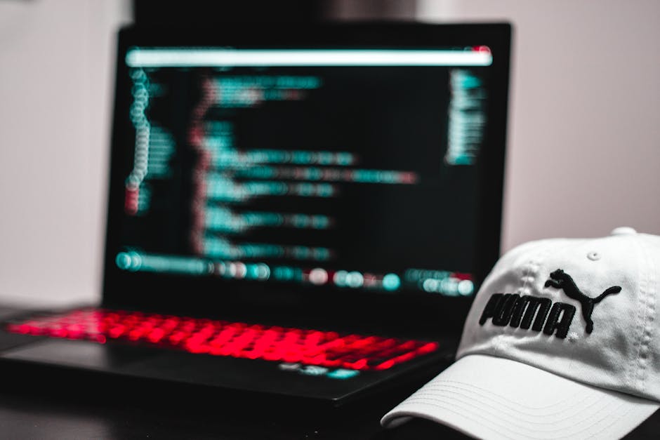 The image features a close-up of a white cap with the Puma logo in black resting on a desk beside a laptop. The laptop's screen is blurred and displays lines of colorful code or data, illuminated with