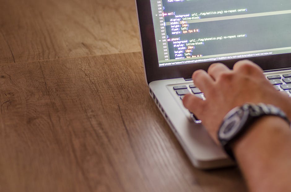 A close-up of a person's hand working on a laptop placed on a wooden desk, with the screen displaying lines of code in a text editor. The individual is wearing a wristwatch with a dark strap, and the